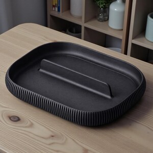 May include: A matte black rectangular tray with a raised central divider and ribbed edges. The tray is made of a solid material and sits on a light-colored wooden surface. The design is minimalist and modern, suitable for home decor.