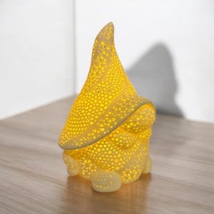 May include: A yellow ceramic gnome-shaped lamp with a textured surface and numerous small holes. The lamp emits a warm, inviting glow. The design features a pointed hat and a rounded base, creating a whimsical and decorative lighting element.