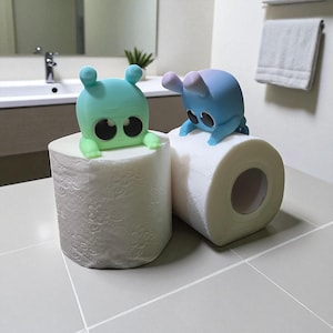 May include: Two rolls of white toilet paper are topped with colorful, cartoonish alien figures. One is teal, the other blue, both with large black eyes. The bathroom setting includes a sink, mirror, and a towel rack.