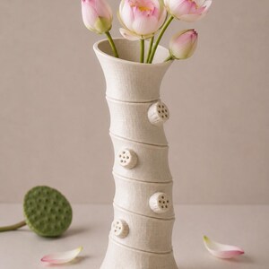 May include: A cream ceramic vase with a textured finish, displaying pink and white lotus flowers. The vase features a distinctive bamboo-style design with circular embellishments. Pink and white petals and a green seed pod are scattered around the vase.