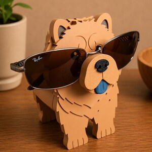 May include: A tan-colored dog-shaped sunglasses holder with a black nose and a blue tongue. The holder is wearing a pair of brown-tinted sunglasses with a silver frame. The sunglasses have the Ray-Ban logo.