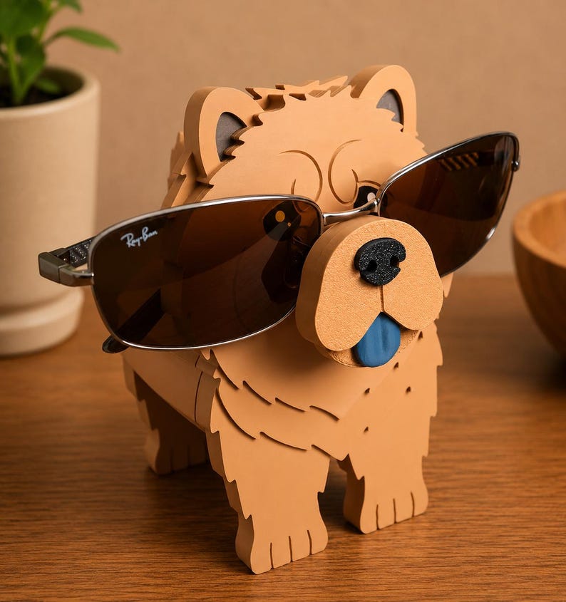May include: A tan-colored dog-shaped sunglasses holder with a black nose and a blue tongue. The holder is wearing a pair of brown-tinted sunglasses with a silver frame. The sunglasses have the Ray-Ban logo.