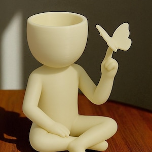May include: A cream-colored figurine of a person in a seated pose, with a butterfly perched on a finger. The figure has a hollow head, designed to hold small plants or items. The figurine is on a wooden surface.