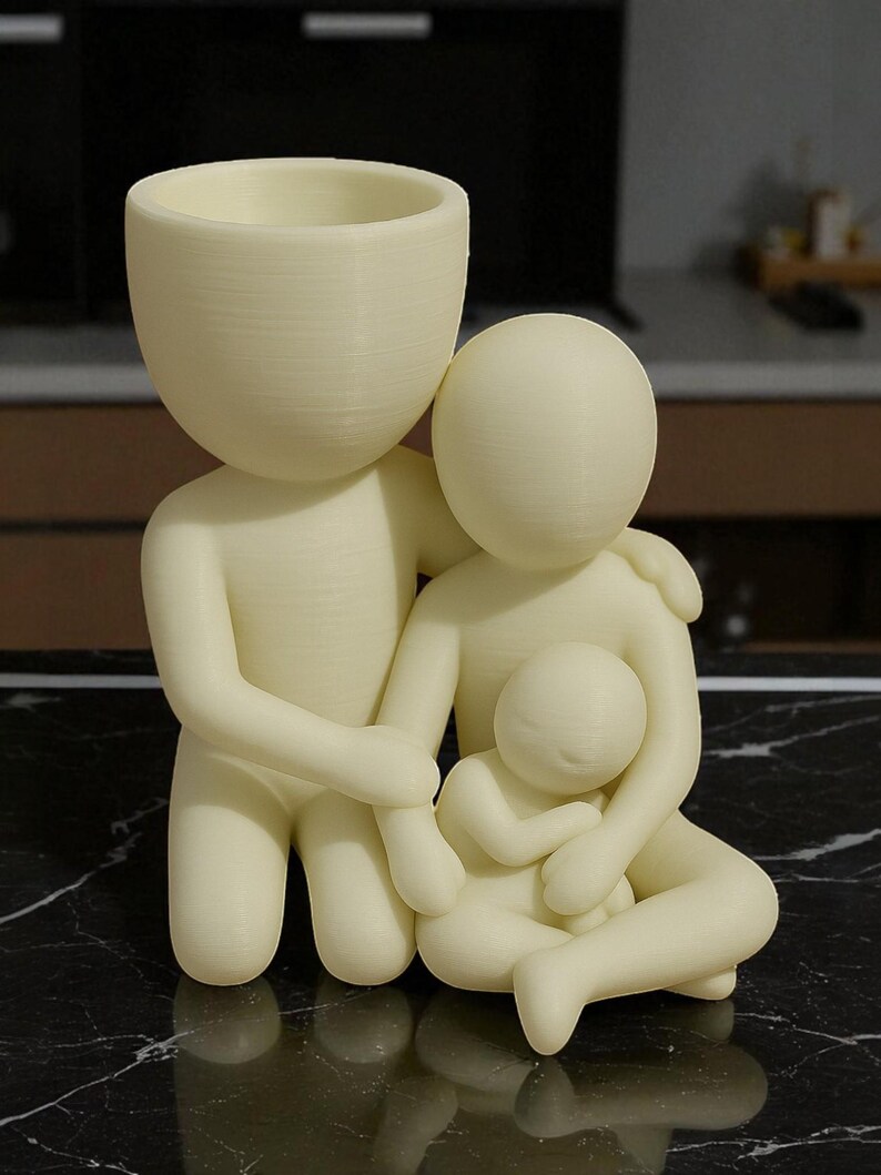 Puede incluir: Figura familiar de color crema. La escultura representa una figura paterna con una cabeza grande y una peque&ntilde;a abertura para maceta, abrazando a otro padre y a un ni&ntilde;o peque&ntilde;o. Las figuras se toman de la mano y se abrazan.
