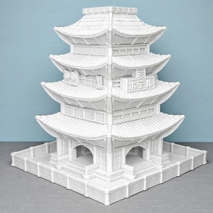 May include: A white, multi-tiered pagoda model with a detailed architectural design. The model features multiple levels with curved roofs and decorative elements. The Japanese text "ホテル" is visible on the side. The pagoda sits on a square base.