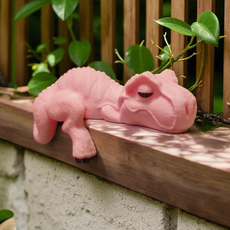 May include: A light pink dinosaur ornament, a Tyrannosaurus Rex, lying on a wooden ledge. The dinosaur's arms and legs are over the edge. The figurine has a smooth texture and a relaxed pose. The ledge is made of wood.