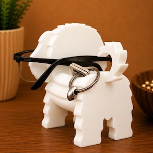 May include: A white, dog-shaped eyeglasses holder with black-framed glasses resting on it. A silver key ring hangs from the dog's tail. The holder is made of a solid, textured material.