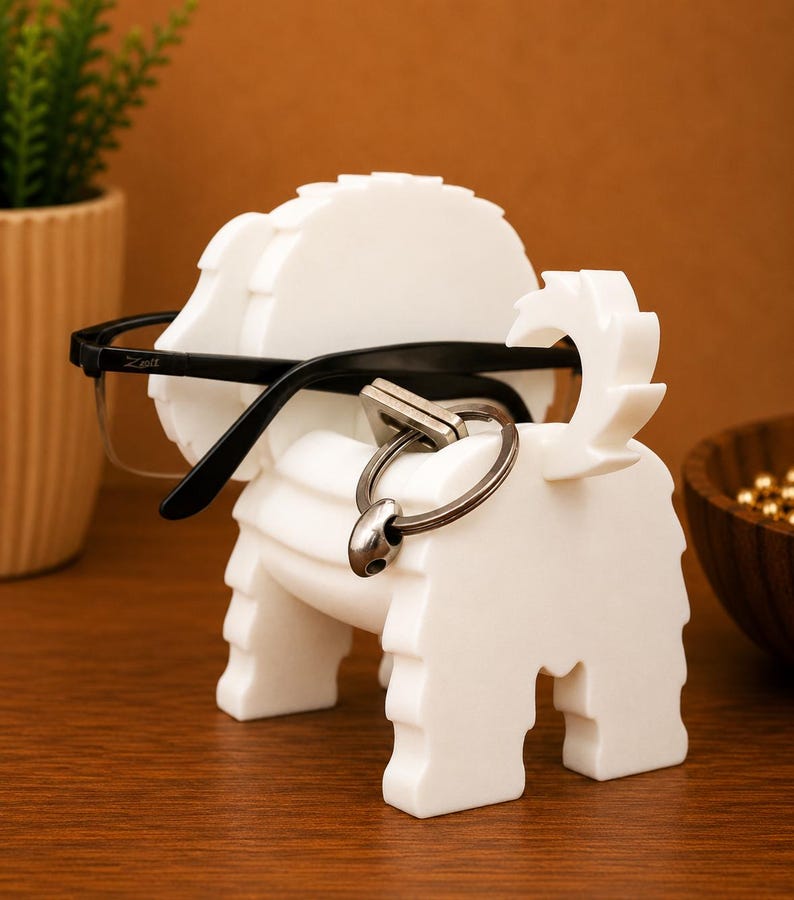 May include: A white, dog-shaped eyeglasses holder with black-framed glasses resting on it. A silver key ring hangs from the dog's tail. The holder is made of a solid, textured material.