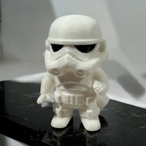 May include: A white, stylized figurine of a Stormtrooper from Star Wars. The figure has a helmet, mask, and armor details. The eyes are black. The figurine is standing on a black surface.