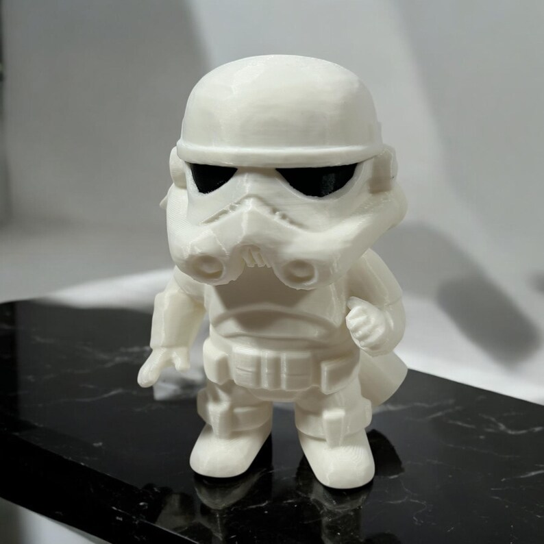 May include: A white, stylized figurine of a Stormtrooper from Star Wars. The figure has a helmet, mask, and armor details. The eyes are black. The figurine is standing on a black surface.