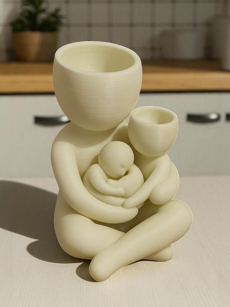 K&ouml;nnte beinhalten: Cremefarbene Keramikskulptur einer Mutterfigur, die zwei Kinder umarmt. Die Skulptur hat eine glatte, matte Oberfl&auml;che und verf&uuml;gt &uuml;ber drei Gef&auml;&szlig;e mit offener Oberseite, die auf einen Pflanztopf oder einen dekorativen Gegenstand hindeuten. Das Design ist minimalistisch und modern.