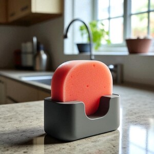 May include: A coral-colored kitchen sponge sits in a dark gray holder. The sponge has a rounded top and is placed in a rectangular holder with a curved opening. The holder is on a countertop in a kitchen setting.