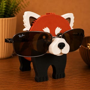 May include: A whimsical red panda-shaped eyeglass holder with a white snout and ears, and black legs. The holder is wearing a pair of Ray-Ban sunglasses with dark lenses. The holder is made of wood and sits on a wooden surface.