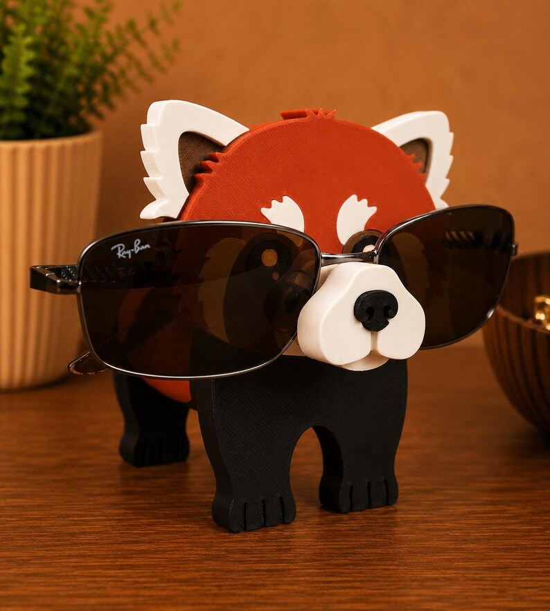 May include: A whimsical red panda-shaped eyeglass holder with a white snout and ears, and black legs. The holder is wearing a pair of Ray-Ban sunglasses with dark lenses. The holder is made of wood and sits on a wooden surface.