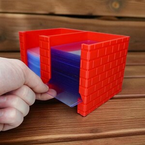 May include: A red, brick-effect card holder with several compartments. The holder contains stacks of blue and clear cards. A hand is pulling a clear card from the holder. The holder is on a wooden surface.