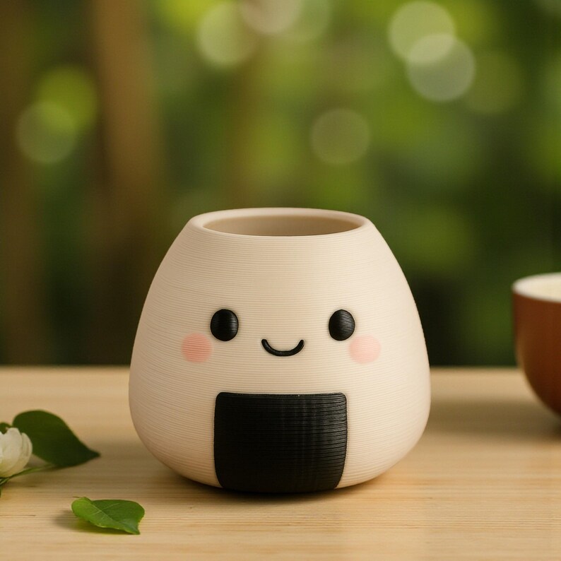 May include: A cream-colored, onigiri-shaped planter with a smiling face, black eyes, and pink cheeks. A black, rectangular seaweed design is at the bottom. The planter is on a wooden surface with a blurred green background.