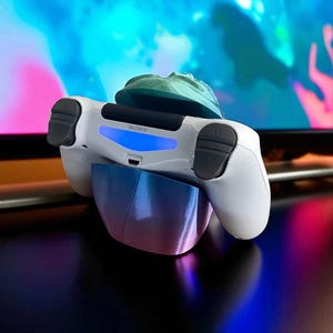 Pu&ograve; includere: Un controller bianco Sony PlayStation 4 appoggiato su un supporto blu e viola. Il controller ha pulsanti neri e una luce blu. Un oggetto verde &egrave; sopra.