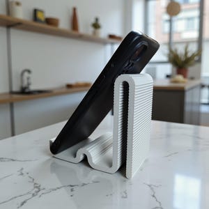 Op de afbeelding: Een witte, getextureerde telefoonstandaard met een golvend ontwerp, die een zwarte smartphone vasthoudt. De standaard is gemaakt van een geribbeld materiaal en is ontworpen om een telefoon rechtop te zetten voor handsfree gebruik. De standaard staat op een wit marmeren oppervlak.