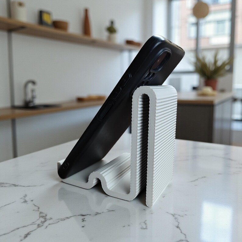Op de afbeelding: Een witte, getextureerde telefoonstandaard met een golvend ontwerp, die een zwarte smartphone vasthoudt. De standaard is gemaakt van een geribbeld materiaal en is ontworpen om een telefoon rechtop te zetten voor handsfree gebruik. De standaard staat op een wit marmeren oppervlak.