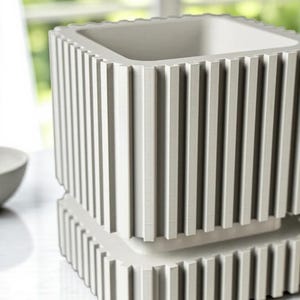 May include: A light gray, square-shaped planter with vertical ribbed detailing. The planter has a two-tiered design, with a base and a main body, both featuring the same ribbed texture. The interior is a smooth, light gray.