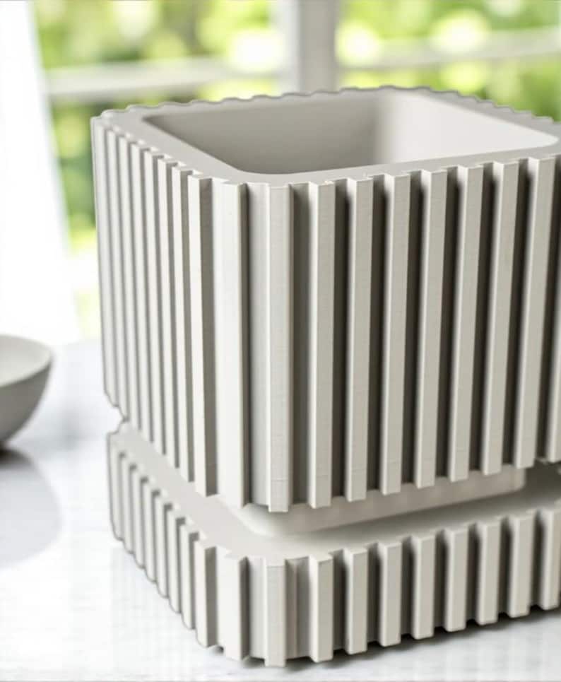 May include: A light gray, square-shaped planter with vertical ribbed detailing. The planter has a two-tiered design, with a base and a main body, both featuring the same ribbed texture. The interior is a smooth, light gray.