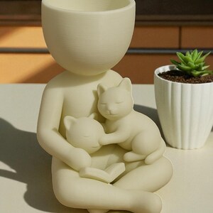 May include: A cream-coloured planter in the shape of a person holding two sleeping cats. The person is seated with legs crossed, holding a book. The top of the person's head is a large, open bowl for planting. A small succulent in a white pot is in the background.