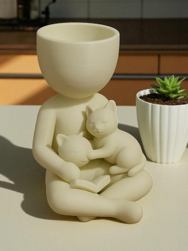 May include: A cream-coloured planter in the shape of a person holding two sleeping cats. The person is seated with legs crossed, holding a book. The top of the person's head is a large, open bowl for planting. A small succulent in a white pot is in the background.