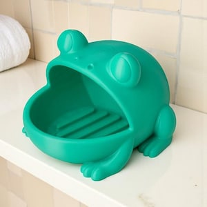 May include: A bright green frog-shaped soap dish. The dish has a wide open mouth for holding soap and features a ridged interior. The frog has large, round eyes and is sitting on a white surface.