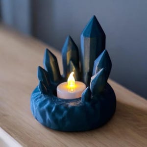 May include: A teal-colored crystal-shaped candle holder with a small, battery-operated candle. The holder features multiple crystal points surrounding the candle, creating a decorative and unique design. The candle emits a warm, yellow glow.