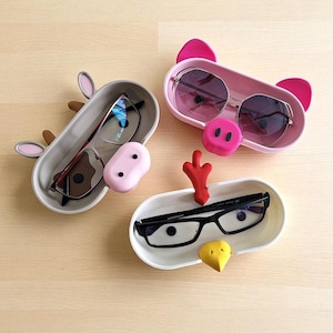 May include: Three whimsical eyeglass holders in the shape of a cow, pig, and chicken. The cow holder is light gray with brown and pink accents. The pig holder is pink, and the chicken holder is white with red and yellow accents. Each holds a pair of eyeglasses.