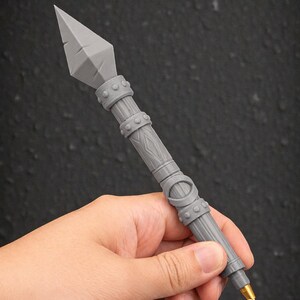 May include: A gray pen with a spearhead design. The pen has a detailed, textured shaft with decorative bands and a gold-colored tip. The pen is held against a dark background.