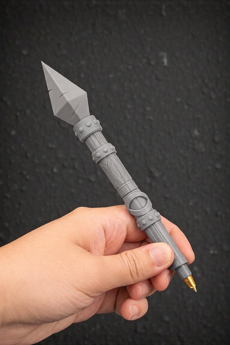 May include: A gray pen with a spearhead design. The pen has a detailed, textured shaft with decorative bands and a gold-colored tip. The pen is held against a dark background.