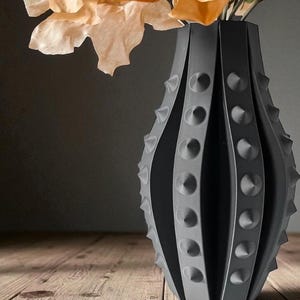 Puede incluir: Un jarr&oacute;n decorativo gris oscuro con un dise&ntilde;o texturizado y puntiagudo. El jarr&oacute;n tiene una forma &uacute;nica con ranuras verticales y detalles en forma de cono en relieve. El jarr&oacute;n contiene flores de color claro, creando un contraste con el jarr&oacute;n oscuro.