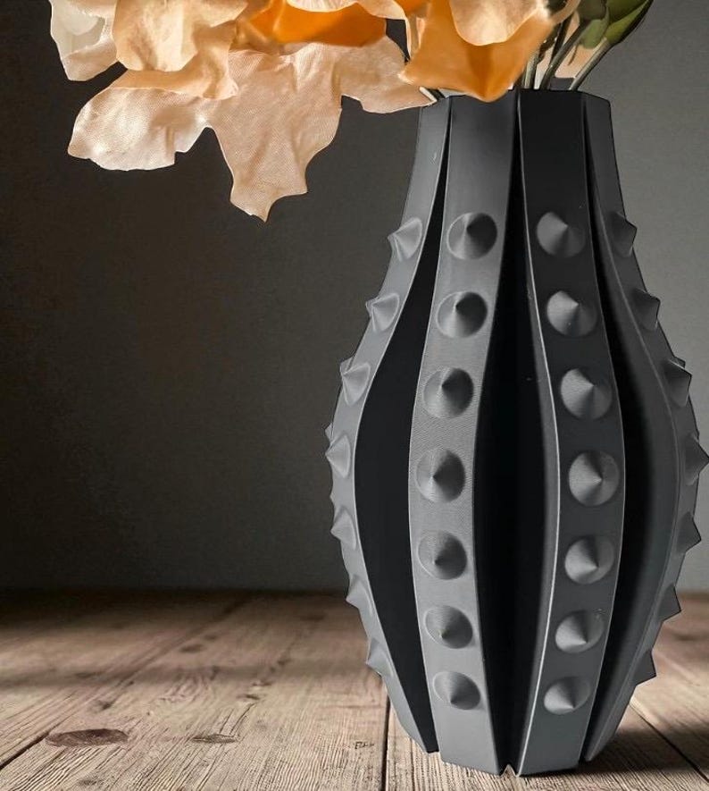 Puede incluir: Un jarr&oacute;n decorativo gris oscuro con un dise&ntilde;o texturizado y puntiagudo. El jarr&oacute;n tiene una forma &uacute;nica con ranuras verticales y detalles en forma de cono en relieve. El jarr&oacute;n contiene flores de color claro, creando un contraste con el jarr&oacute;n oscuro.