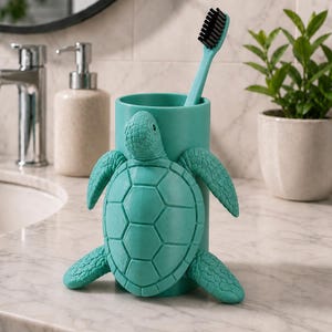 May include: A teal toothbrush holder shaped like a turtle, with a matching teal toothbrush. The turtle has detailed flippers and a textured shell. The holder is on a marble countertop, with a sink, faucet, and soap dispenser in the background.