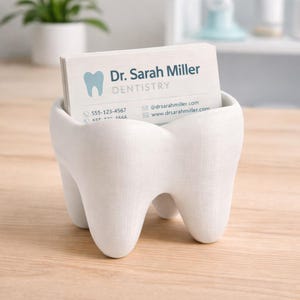 May include: A white, tooth-shaped business card holder containing a stack of cards. The cards show "Dr. Sarah Miller, Dentistry" and contact details. The holder sits on a light-coloured wooden surface.