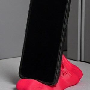 May include: A black smartphone in a protective case rests on a bright pink, abstract-shaped phone stand. The stand is made of a smooth material and is designed to hold the phone upright.