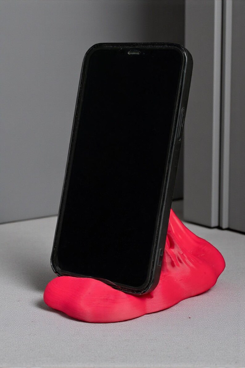 May include: A black smartphone in a protective case rests on a bright pink, abstract-shaped phone stand. The stand is made of a smooth material and is designed to hold the phone upright.