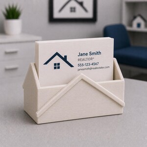 Pu&ograve; includere: Un portacarte beige chiaro a forma di casa, contenente una pila di biglietti da visita bianchi. Le carte mostrano un logo a forma di casa blu navy e il nome "Jane Smith, REALTOR&reg;" con le informazioni di contatto. Una piccola pianta e un'opera d'arte sono sullo sfondo.