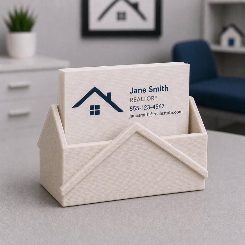 Pu&ograve; includere: Un portacarte beige chiaro a forma di casa, contenente una pila di biglietti da visita bianchi. Le carte mostrano un logo a forma di casa blu navy e il nome "Jane Smith, REALTOR&reg;" con le informazioni di contatto. Una piccola pianta e un'opera d'arte sono sullo sfondo.