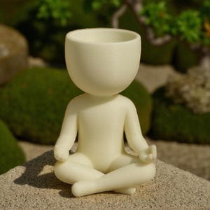 Puede incluir: Figura de color crema de una persona en posición de yoga sentado. La cabeza tiene un diseño en forma de copa. La figura está sentada sobre una superficie de piedra con un fondo verde borroso.