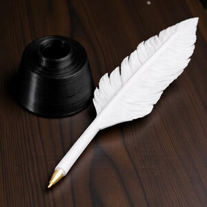 May include: A white feather pen with a gold-colored tip rests on a dark wooden surface next to a black inkwell. The pen is designed to resemble a feather, with detailed feathering. The inkwell is a simple, cylindrical shape.