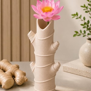 Op de afbeelding: Een beige keramische vaas in de vorm van een gemberwortel, met een roze lotusbloem met een gele kern. De vaas heeft een uniek, getextureerd ontwerp. Gemberwortel is zichtbaar op de voorgrond. Een kleine vaas met groen is op de achtergrond.