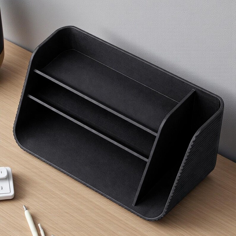 May include: A black desk organizer with multiple compartments. The organizer has a rectangular base with rounded corners and a textured exterior. It features vertical dividers to separate items, providing storage for office supplies.