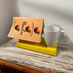 May include: A book titled "Peaches" with peach illustrations is propped up on a yellow wooden stand. A white mug sits next to the book. The book and mug are on a wooden surface.