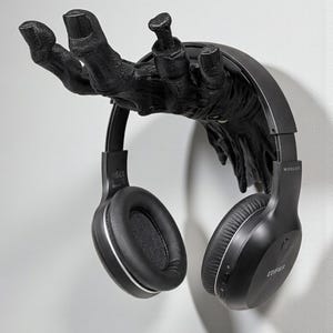 K&ouml;nnte beinhalten: Ein schwarzer, an der Wand befestigter Kopfh&ouml;rerhalter in Form einer Hand. Der Halter tr&auml;gt ein Paar schwarze kabellose Kopfh&ouml;rer mit silbernen Akzenten. Ein einzigartiges und funktionales Design.