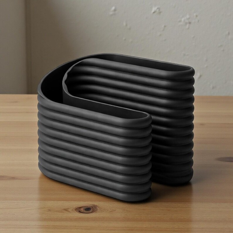 May include: A modern, black, sculptural vase with a unique, layered design. The vase has a curved, rectangular shape with horizontal ridges, creating a textured surface. The vase is made of a matte black material and sits on a wooden surface.