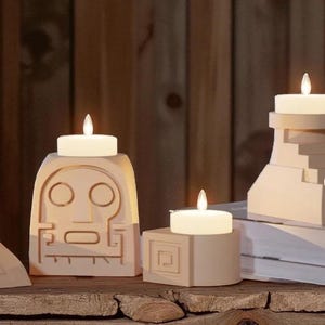 May include: Beige candle holders with lit white candles. One holder has a face design, another is a cube with a geometric pattern, and a third is a stepped design. The holders sit on a wooden surface against a wooden background.