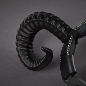 May include: Black headphones featuring a curved, textured horn-like element. The headphones have a padded headband and earcups, and a zip detail. The design is dark and edgy, with a focus on the horn-shaped feature.