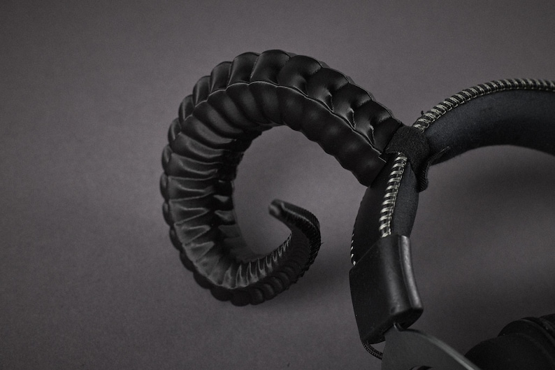 May include: Black headphones featuring a curved, textured horn-like element. The headphones have a padded headband and earcups, and a zip detail. The design is dark and edgy, with a focus on the horn-shaped feature.